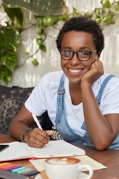 Glad black female author wears transparent glasses and piercing