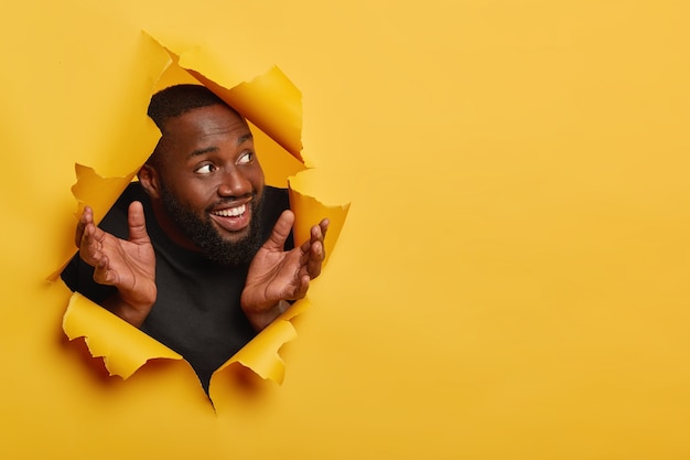 Glad black man spreads palms sideways, looks happily aside, notices something pleasant, has thick bristle, white teeth, poses in torn hole of yellow background