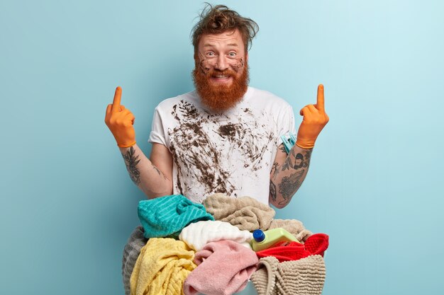 Glad blue eyed redhead man with thick bristle, has dirty white t shirt, wears rubber gloves, shows middle finger with both hands, stands near basin of laundry, isolated over blue wall.