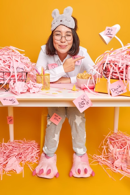 Free photo glad busy female office worker indicates at pile of cut paper shows mess at desktop wears sleepmask and pajama satisfied to finish work isolated on yellow makes notes on stickers