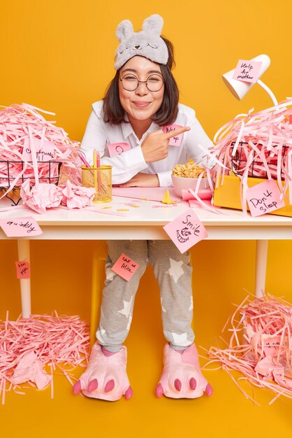 glad busy female office worker indicates at pile of cut paper shows mess at desktop wears sleepmask and pajama satisfied to finish work isolated on yellow makes notes on stickers