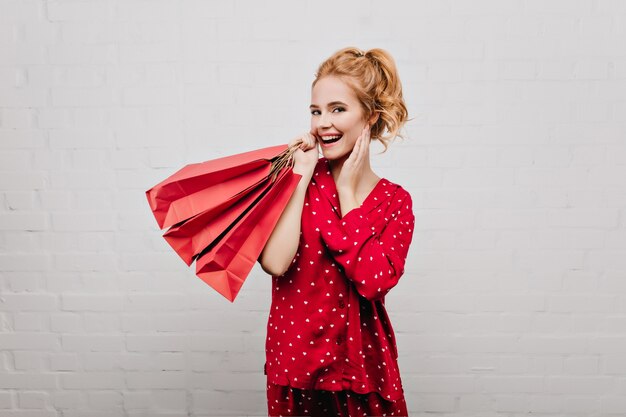 Glad caucasian woman wears cotton red pajamas posing with paper bags. Indoor portrait of emotional blonde girl with gifts isolated on light wall