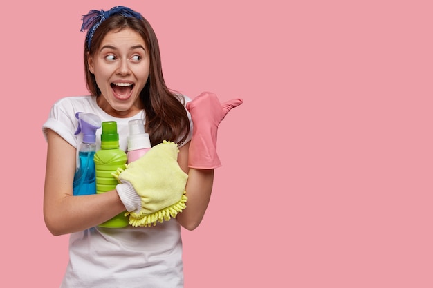 Glad Caucasian woman with positive expression, wears rubber gloves, points aside with thumb, holds spray and detergent in hands