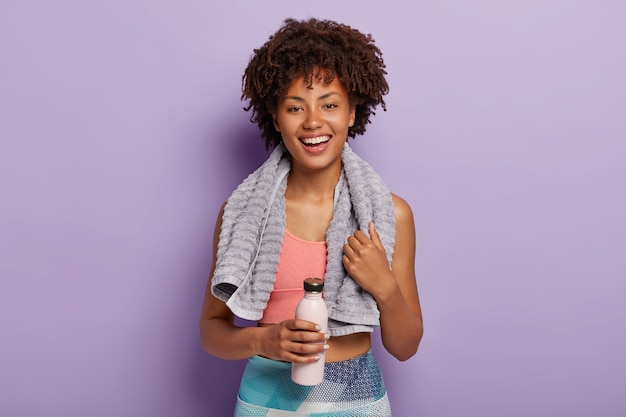 Free photo glad charming fitness woman drinks cold water, being thirsty after running, has towel on neck