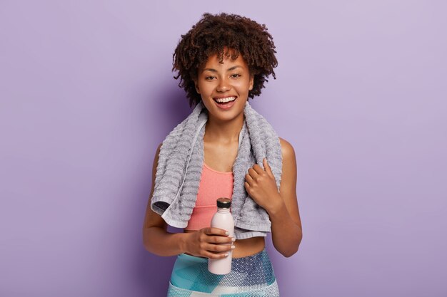 Обаятельная фитнес-женщина пьет холодную воду, хочет пить после пробежки, с полотенцем на шее