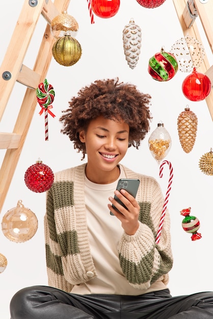 Free photo glad charming millennial girl with afro hair scrolls social media via smartphone sits relaxed indoor takes break after decorating home for coming winter holidays surfs internet makes shopping online