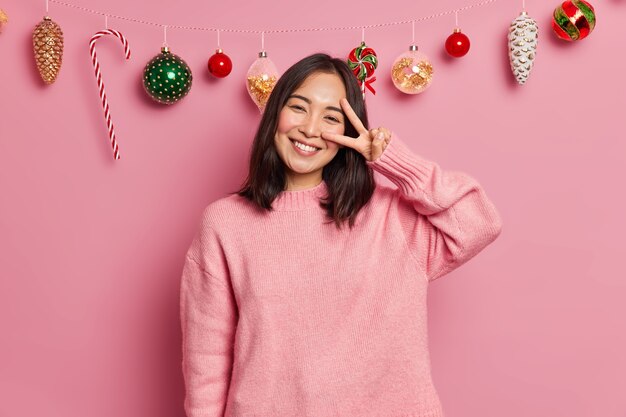 Glad charming woman with toothy smile dark hair makes peace gesture expresses positive emotions dressed in casual sweater has festive mood poses 