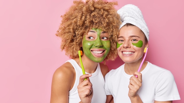 Glad cheerful women apply green beauty mask and hydrogel patches hold toothbrushes dressed in casual t shirts isolated over pink background with blank space for your advertisement Skin care
