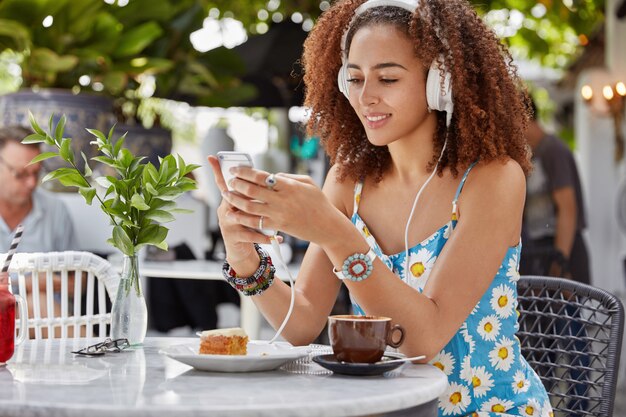 嬉しい集中アフリカ系アメリカ人の女性は、スマートフォンとヘッドフォンに接続されているプレイリストからお気に入りの曲を楽しんで、屋外のコーヒーショップに座っています。
