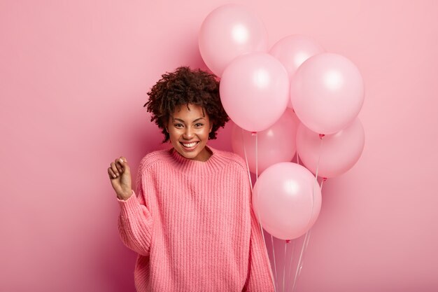 기쁜 곱슬 여성 모델은 주먹을 쥐고, 특대 점퍼를 착용하고, 공기 풍선을 들고, 생일 축하에 참석하게되어 행복하고, 벽에 한 톤의 분홍색 스웨터를 입습니다.