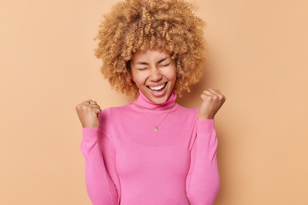 Free photo glad curly haired woman clenches fists with triumpg feels like winner celebrates triumph wears casual pink turtleneck isolated over beige background rejoices victory achieves goals. yes i did it