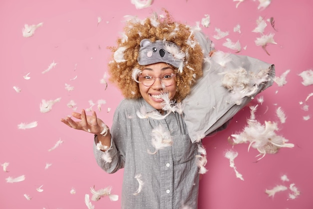 Free photo glad curly haired young woman has fun dressed in pajama holds pillow poses against pink background with feathers flying around in air overjoyed female model has pillow fight after awakening