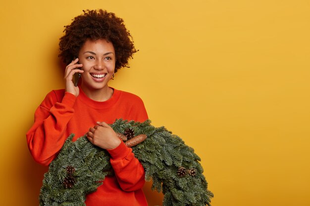 glad curly woman enjoys telephone conversation, discusses Christmas preparation with friend, holds fir handmade wreath with pine cones, stands against yellow background