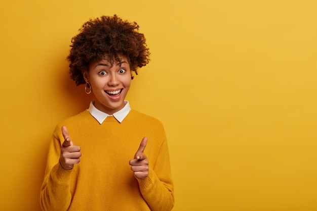 Glad curly woman points both index fingers at you, shows gun sign