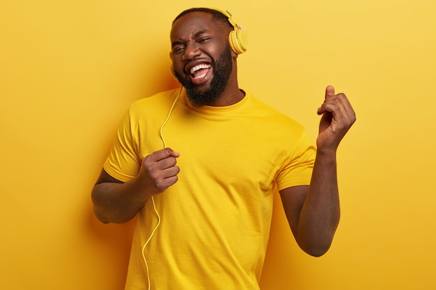 Glad dark skinned plump man dances, makes movements to music, has modern stereo headphones, smiles positively, being in high spirit. Everything in yellow color. Carefree guy listens lively music