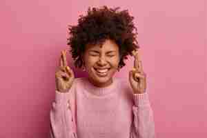 Free photo glad dark skinned woman stands with crossed fingers, anticipates important news, gestures indoor, has curly hair, wears casual jumper, makes sweet desire, isolated over pink wall. body language