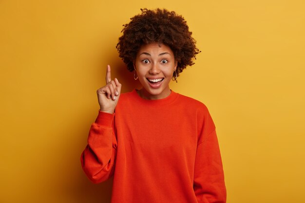 Glad dark skinned woman with Afro haircut recommends awesome product, points up satisfied, shows you perfect promo, dresssed in red sweater, isolated over yellow studio wall