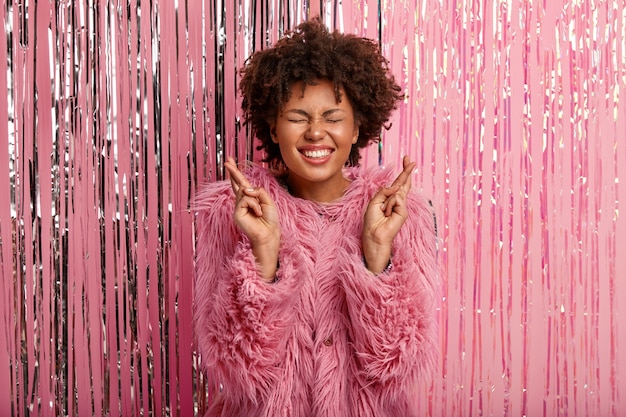 Free photo glad dark skinned young female with broad smile, wears fashionable pink coat, crosses fingers