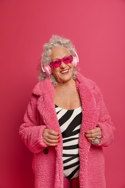 Glad elderly woman wears fashionable coat, follows fashion trends, enjoys music