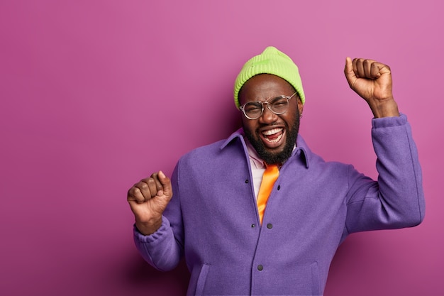 Glad enthusiastic bearded man keeps hands up, celebrates triumph, wears stylish clothes