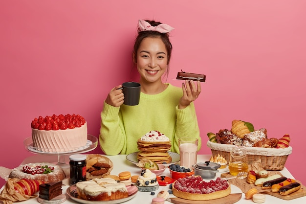 Glad ethnic woman holds piece of chocolate cake, drinks tea with dessert, celebrates holiday at home with delicious sweet food, gets pleasure and enjoyment from unforgettable taste.