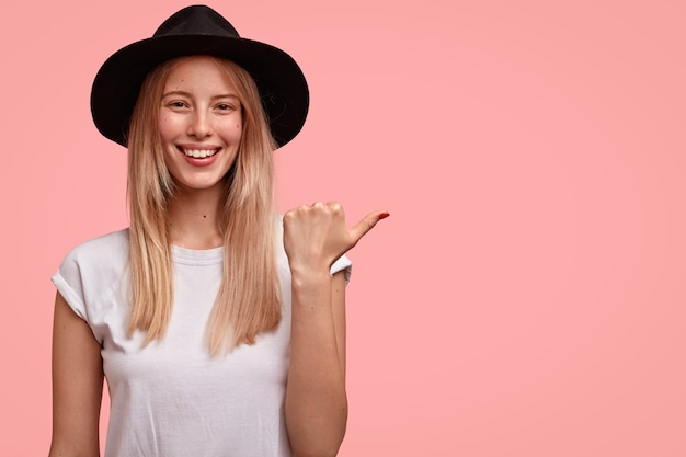 Glad European female has broad smile, wears elegant hat, points with thumb aside, shows direction to stranger, looks freindly, isolated over pink wall