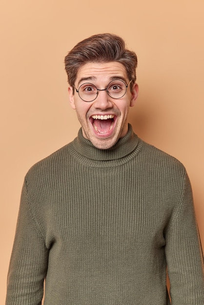 Glad European man exclaims loudly keeps mouth opened reacts on something awesome being very emotionally dressed casually isolated over beige background. Human reactions and emotions concept.