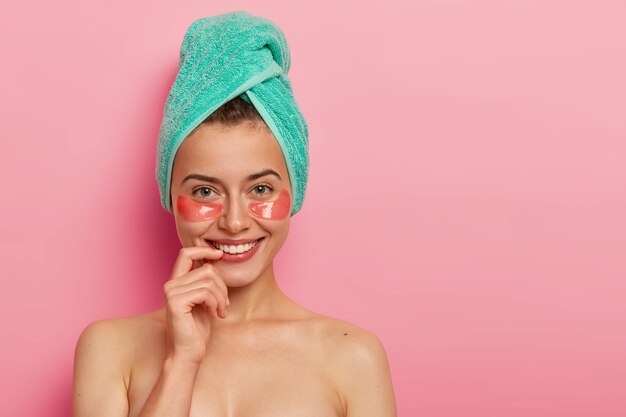 Glad European woman takes care of delicate skin around eyes, applies collagen patches, wears minimal makeup, wrapped bath towel on head, stands naked against pink background.