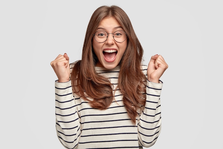 Free Photo glad european woman wears spectacles, clenches fists in excitement, wears round spectacles and casual sweater, exclaims with happiness, has dark hair and pleasant appearance, models over white wall