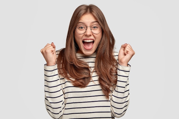 Glad European woman wears spectacles, clenches fists in excitement, wears round spectacles and casual sweater, exclaims with happiness, has dark hair and pleasant appearance, models over white wall