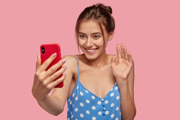 Радостная европейская женщина с приятной улыбкой, машет рукой мобильного телефона, одетая в платье в горошек