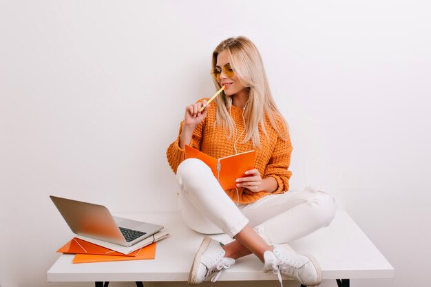 Glad fair-haired woman in trendy outfit doing her job in office, holding notebook and pen