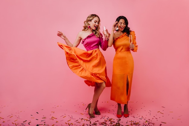 Glad fairhaired girl dancing in studio indoor full length shot of two ladies fooling around on pink background