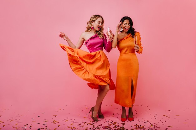 スタジオで踊っているうれしい金髪の女の子ピンクの背景に浮気している2人の女性の屋内全身ショット
