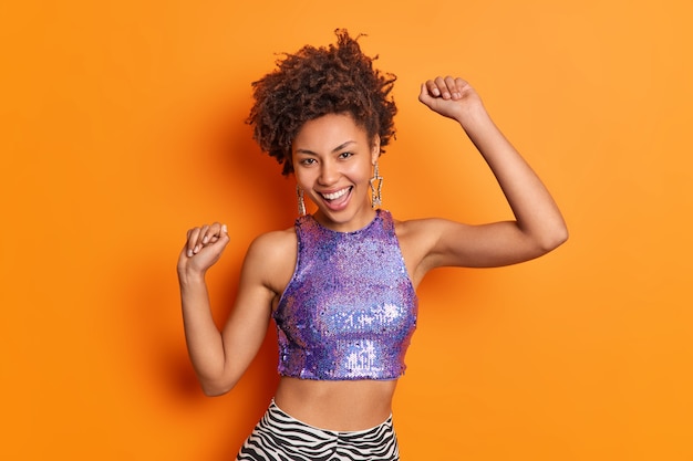 곱슬 머리를 가진 기쁜 유행 아프리카 계 미국인 여자는 보라색 빛나는 탑을 입은 평온한 춤을 생생한 오렌지 벽 위에 고립 된 팔을 올립니다.
