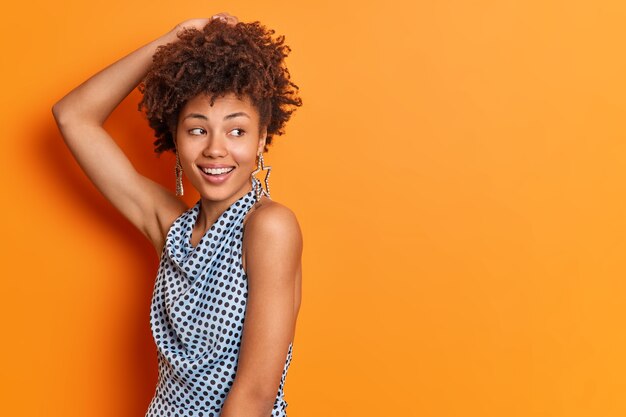 Glad fashionable lady stands sideways keeps hand on head dressed in polka dot blouse smiles broadly isolated over orange wall