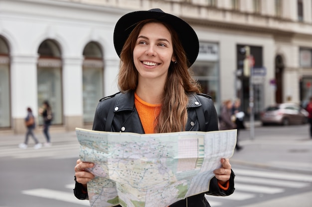 Glad female holds paper map, visits city, looks around, tries find new destination, wears black hat