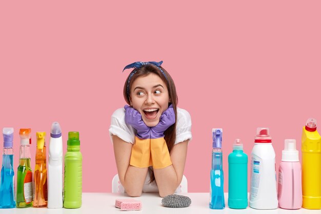 嬉しい女性用務員は、手をあごの下に置き、幸せそうに見え、ヘッドバンドとカジュアルなTシャツを着て、洗剤とスポンジを使って掃除し、ピンクの壁に隔離します。家庭のコンセプト。