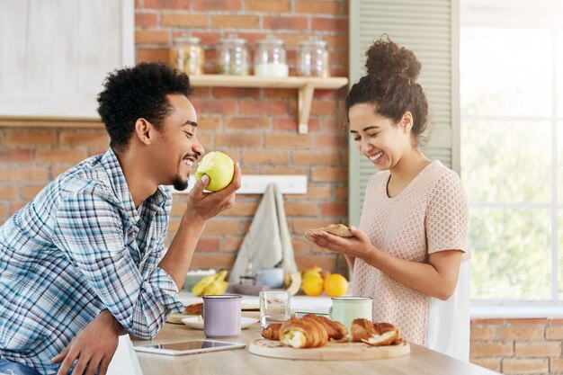 Glad female makes sandwich, talks with husband who sits opposite her, eats apple. Family couple spends free time together,