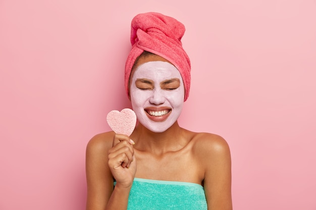 Glad female model smiles broadly, shows white teeth, holds heart shaped cosmetic sponge, keeps eyes shut from pleasure