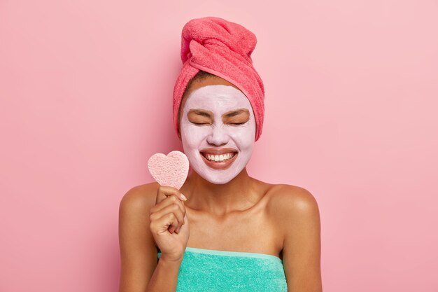 Glad female model smiles broadly, shows white teeth, holds heart shaped cosmetic sponge, keeps eyes shut from pleasure