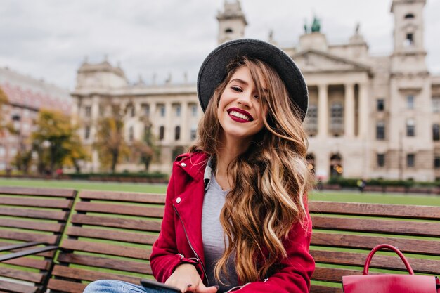 Glad female model with blonde wavy hair sitting on wooden bench in weekend day