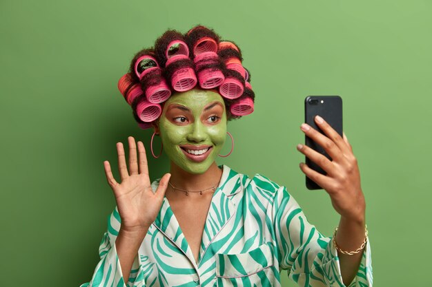 Glad female model with green facial mask, waves palm and says hello to friend, has video conference via modern smart phone, wears hair curlers for making perfect haircut, dressed in casual clothing