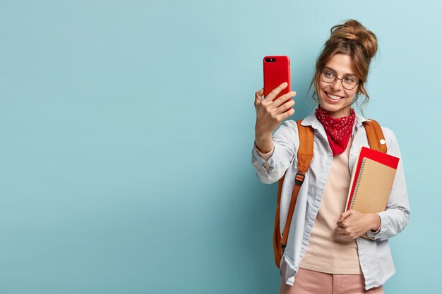 Glad female model with positive smile, makes selfie portrait on her cell phone