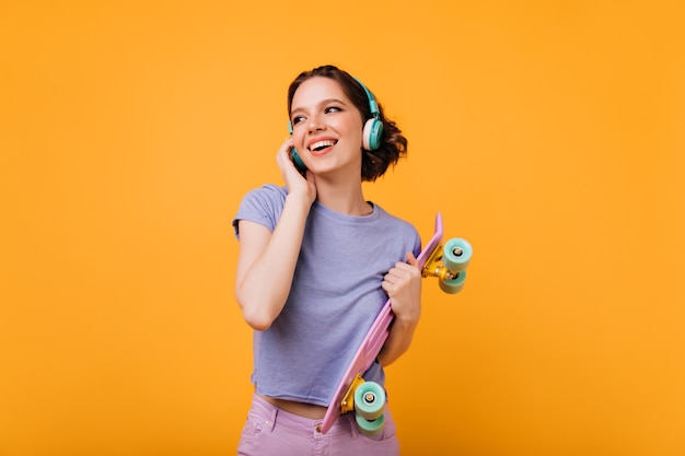 Glad female skateboarder listening favorite song. Indoor shot of blissful curly girl with wavy hairstyle holding her pink longboard.