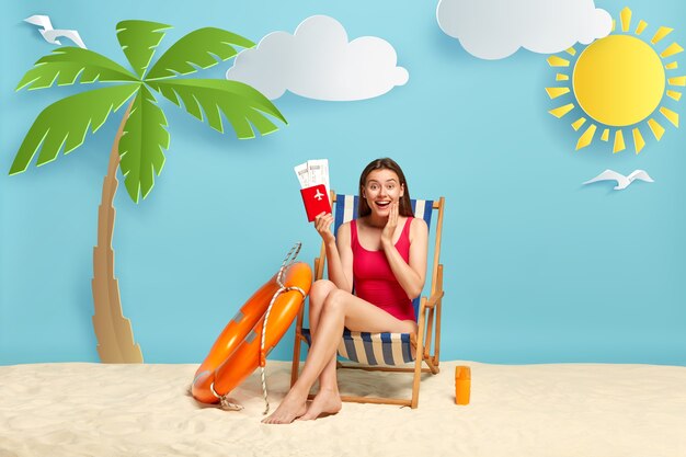 Glad female traveler sits on beach chair, holds passport with flying tickets