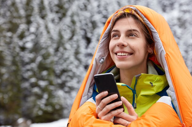Glad female traveler warms in sleeping bag, poses on top over mountain covered with snow, holds modern cell phone
