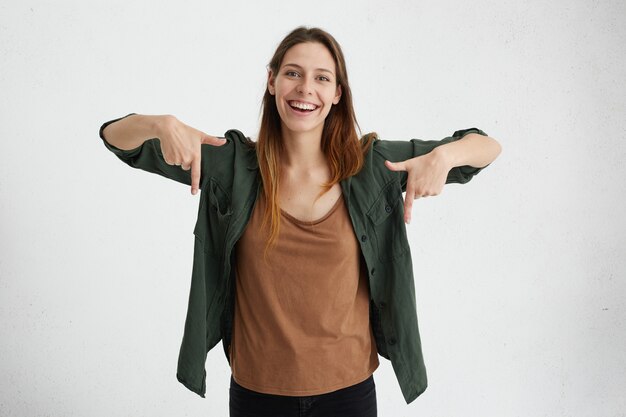 Glad female with oval face, dark straight hair wearing green jacket and brown shirt pointing with her index fingers down having cheerful look