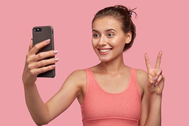 glad female with toothy smile, combed hair, has sporty body, makes peace sign or V gesture of cell phone, poses for making selfie, isolated over pink wall. Video call