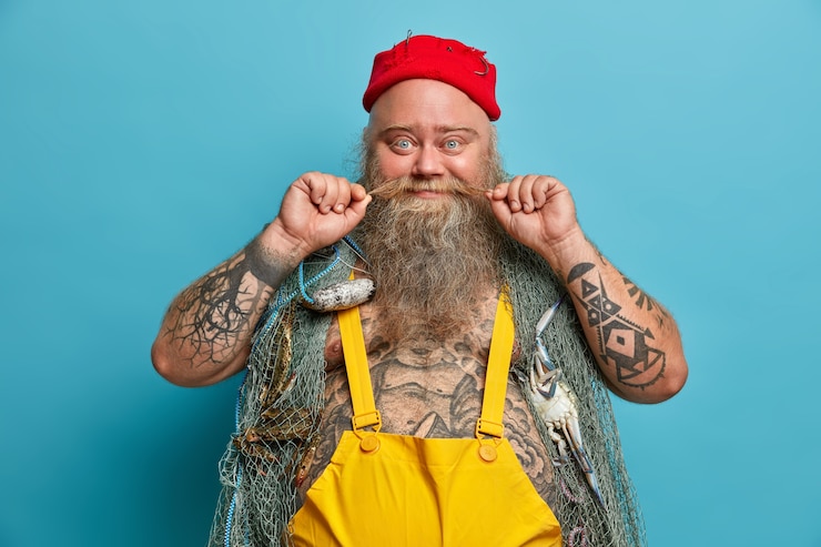 Free Photo glad fisherman curls mustache, has thick beard, carries fishing net on shoulders, spends free time for hobby and soul, wears red hat and overalls, has tattoed body